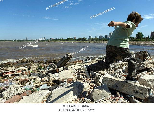 Boy running on shore littered with debris, city in background