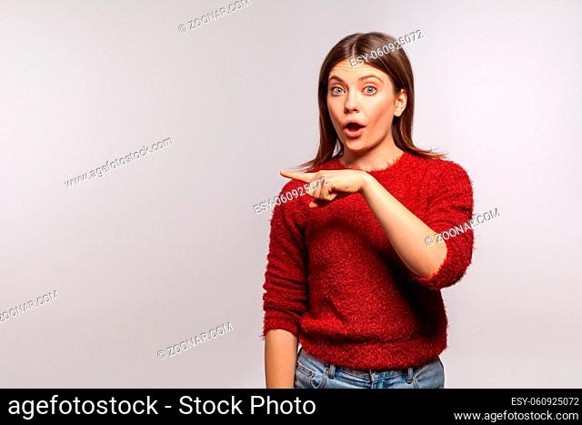 Wow, look at advertisement. Portrait of surprised shocked girl in shaggy sweater pointing empty space, showing blank wall for commercial idea, product image