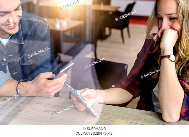 Male and female students looking at smartphone in cafe window seat, view through window