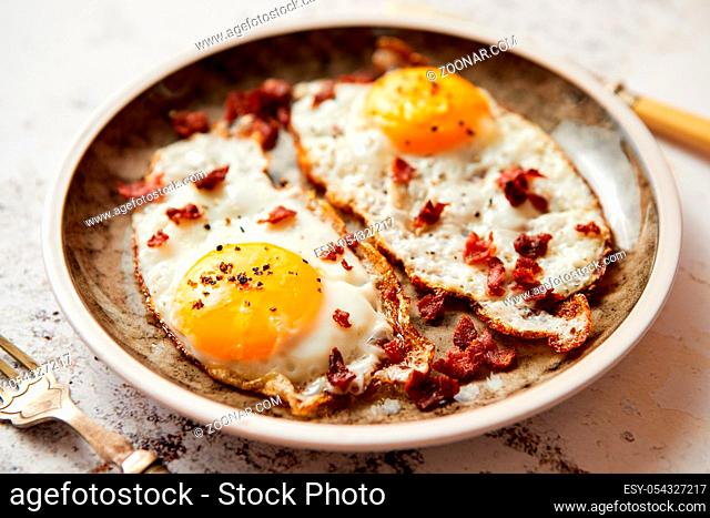 Two fresh fried eggs with crunchy crisp bacon served on rustic plate. Fork and knife on sides. Top view