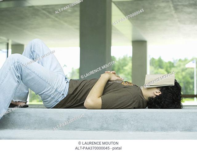 Male college student lying on ground with book covering face