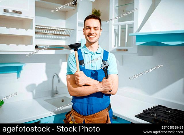 Cheerful male plumber in uniform holds wrench and plunger in the kitchen. Handyman with toolbag repair sink, sanitary equipment service at home