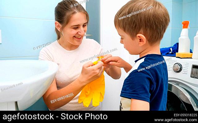 Smiling young mother helping little son putting on yellow rubber gloves before doing housework in bathroom. Children helping parent with chores and daily...