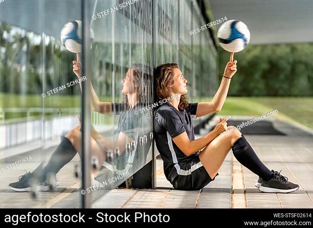 Female soccer player spinning ball on finger while sitting by glass wall