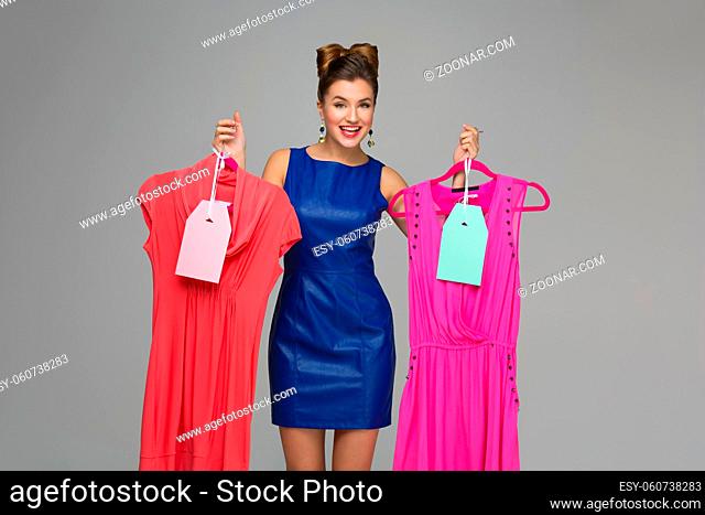 Beautiful happy woman holding two dresses on hangers. Price tags. Over grey background. Copy space