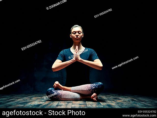 Young sporty woman practicing yoga doing Ardha Padmasana exercise with namaste in dark room. Lotus pose, meditation concept