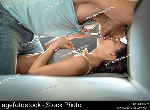 Passionate kissing couple with closed eyes on the sofa. Girl in a gray top holds a flower in left hand. Guy in light sleeveless and blue jeans stands on the...
