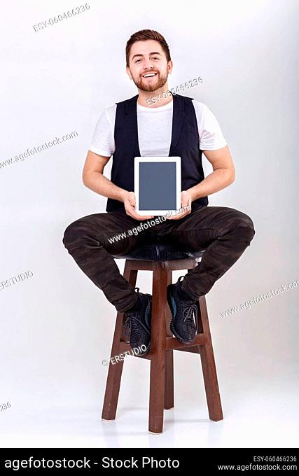 young handsome smilling brunette man with beard in white shirt and black waistcoat holding tablet and sits on the chair on gray background