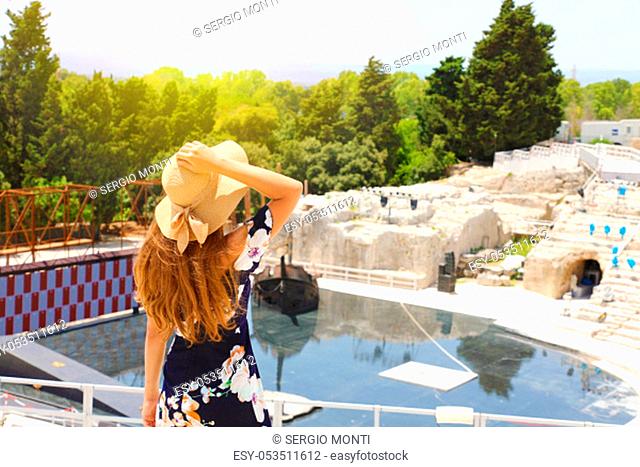 Beautiful elegant woman with hat and flowered dress in Syracuse Greek theater in Sicily, Italy, back view outdoors