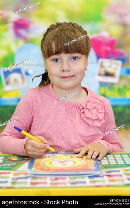 A cute preschool girl is sitting at a school desk holding a pen and looking at the camera