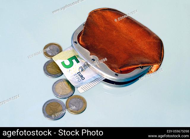 Leicht geoeffnete altmodische Geldboerse aus braunem Leder, aus der ein Fuenf-Euro-Schein herausragt, davor liegen fuenf einzelne Muenzen