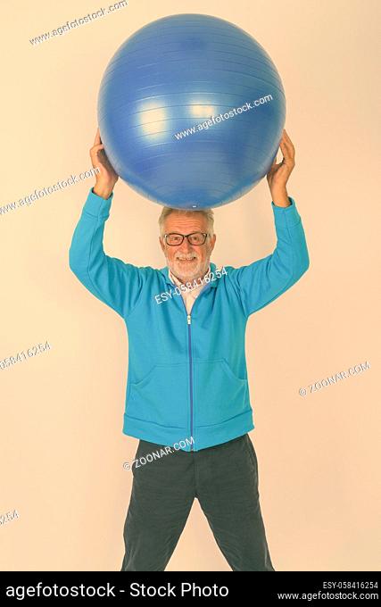 Studio shot of happy senior bearded man smiling and standing while holding gym ball on top of head ready for gym against white background