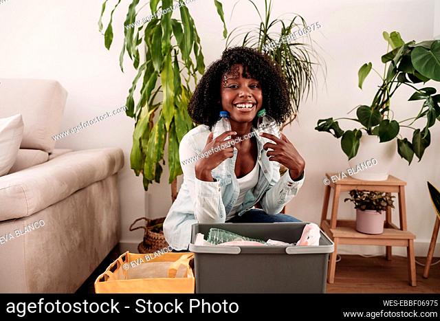 Smiling woman holding plastic bottles crouching at home