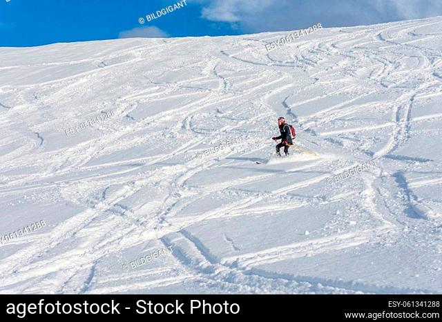 Woman with brown hair, wearing ski wear and a bagpack skiing down a beautiful snow hill with plenty of tracks on the snow, view from the side from far away