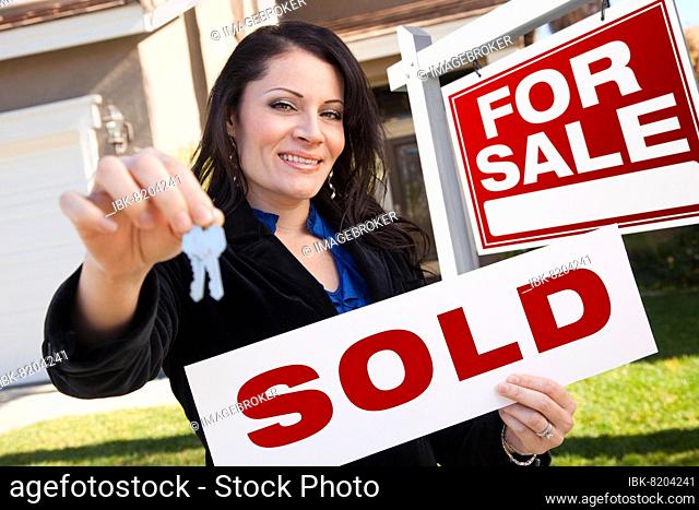 Happy attractive hispanic woman holding sold real estate sign and keys in front for sale real estate sign and house