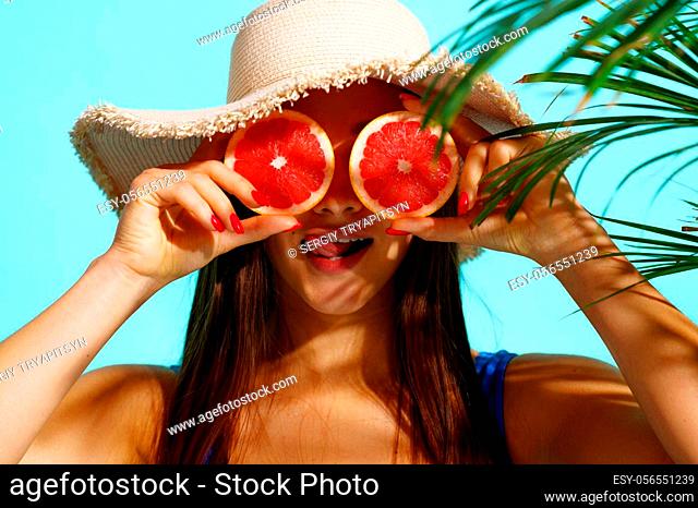 Pretty woman in swimsuit poses with fruits in studio, cyan background. Girl in swimwear ready to take a tan. Model with slim body in swimming underwear