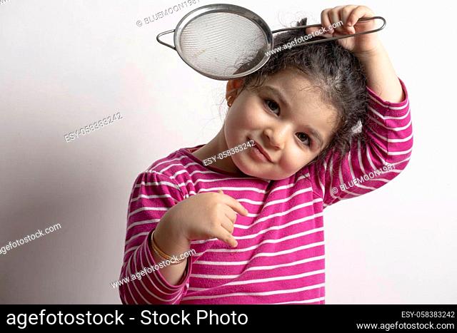 Little girl playing with a strainer in front of her head looking to the camera, childhood memories cooking experience