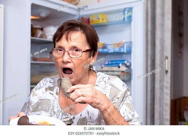 Senior woman going to eat pork liver sausage while standing in front of the open fridge in the kitchen