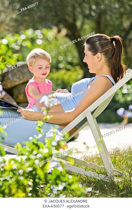 Daughter touching her pregnant mothers belly while she is relaxing in the garden on a lounge chair