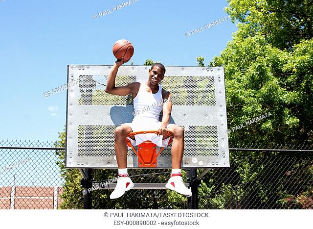 Happy smiling handsome sporty African-American male basketball player dressed in white and holding his ball with one hand outdoor on a summer day in a...
