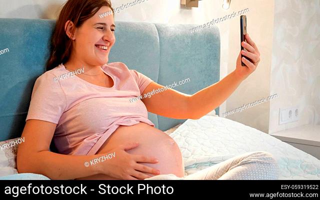 Smiling pregnant woman waiting for baby sitting in bed and calling friend or doctor on video call conference. Pregnant woman using smartphone
