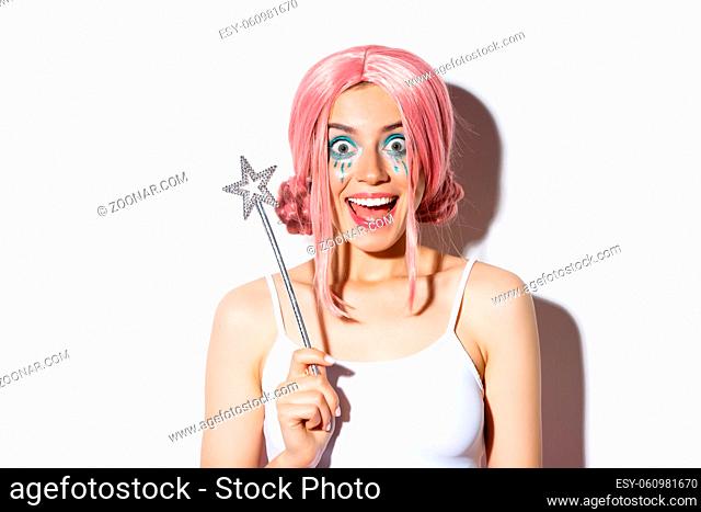 Close-up of surprised happy girl in halloween costume of fairy, holding magic wand and smiling amazed at camera, standing over white background