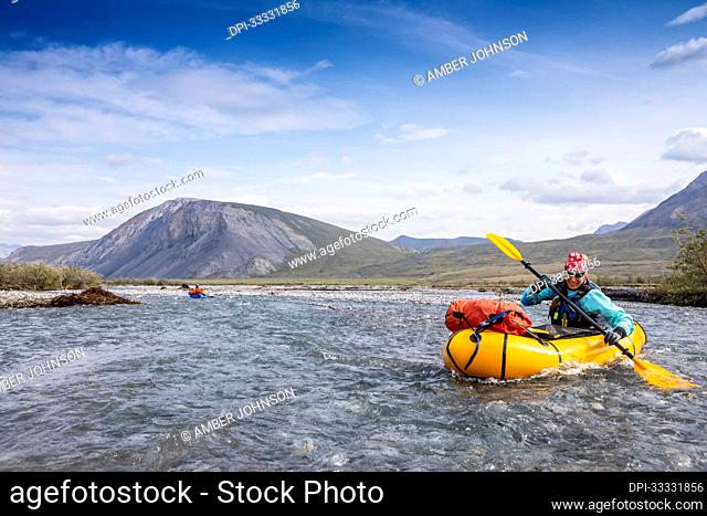Caucasian woman in her 40's smiles as she digs deep with her paddle into the river, guiding her yellow packraft down the crystal clear Marsh Fork River