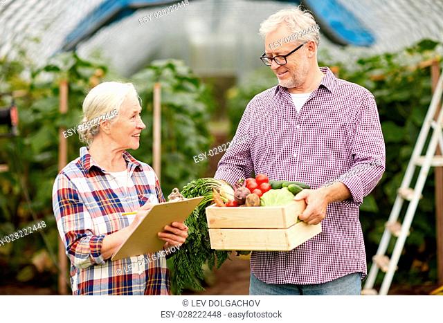 farming, gardening, agriculture, harvesting and people concept - senior couple with box of vegetables and clipboard at farm greenhouse