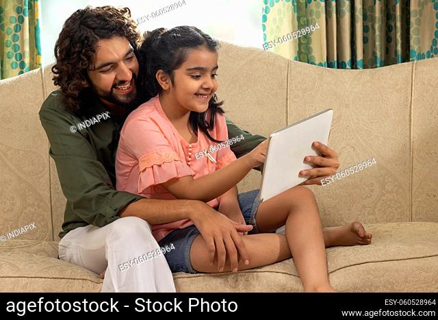 Father and daughter playinggames on tablet together while sitting on sofa at home