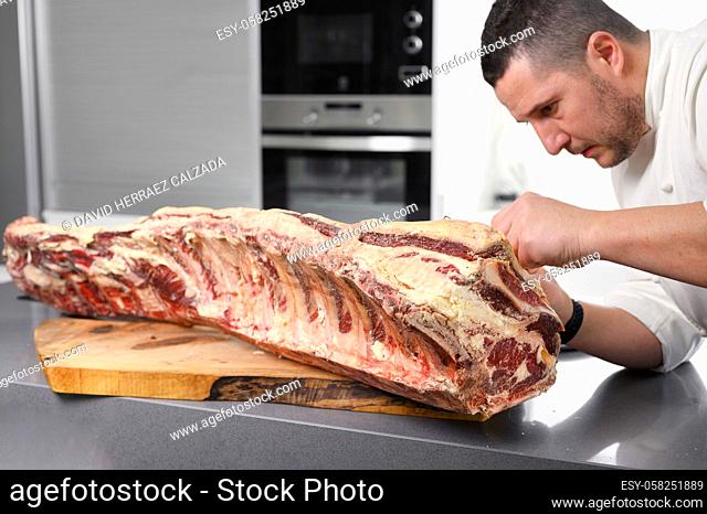 Chef is preparing prime rib steaks from a large prime beef rib roast with multiple bones. The meat is on a wooden board which is sitting on a modern kitchen...