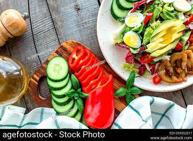 Healthy eating and Mediterranean diet concept flat lay. Plate with lettuce salad leaves, quail egg, avocado, tomato, cutting board with cucumber, pepper, basil