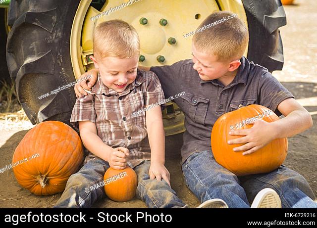 Two boys sitting against a tractor-tire holding pumpkins and talking in rustic setting