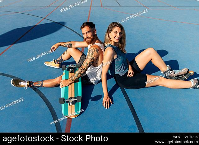 Smiling young couple sitting back to back at basketball court on sunny day