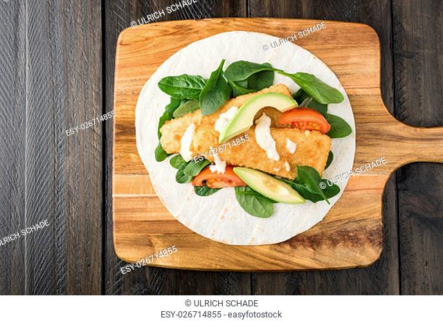 Crumbed fish fillet burrito with avocado and tomato serves on wooden cheese platter with rustic background