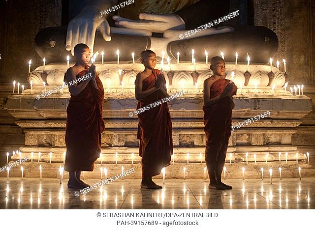 Three young Buddhist monks are standing with a candle in a temple in front of a Buddha statue in Bagan, Myanmar, on 01.04.2013