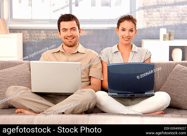 Happy couple sitting on sofa at home, browsing internet on separate laptops, smiling, having fun
