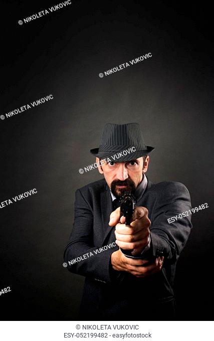 Old-fashioned gangster in suit and hat is aiming with pistol on dark background. Retro and vintage style