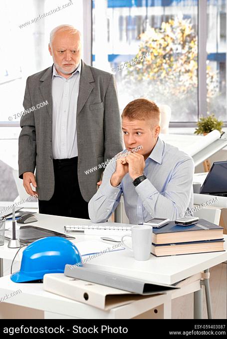 Scared young office worker man sitting at desk, angry senior executive standing