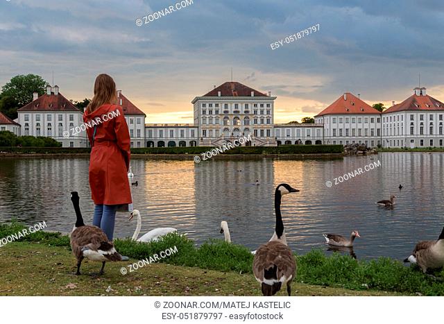 Dramatic scenery of Nymphenburg palace in Munich Germany. Sunset after the sorm. Woman feeding white swans and duks swimming in pond in front of the palace