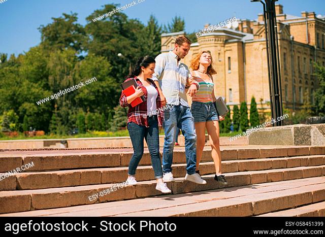 Happy students are walking together on campus. One man with two girls. Students between lectures getting sown the stairs in a park