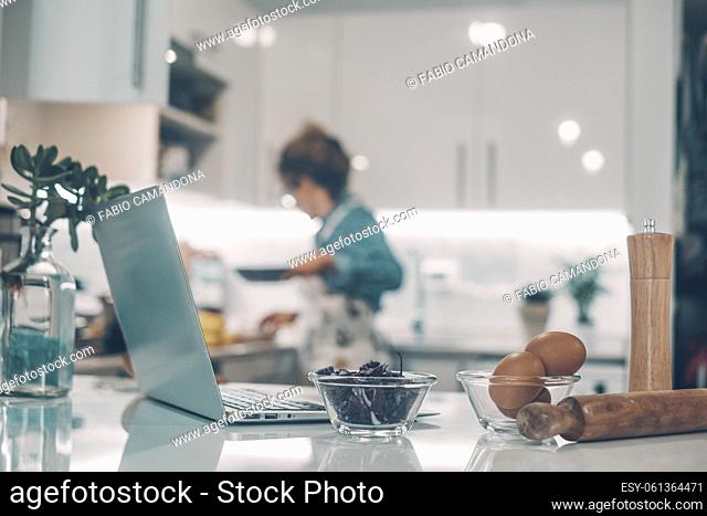 Foreground focus on laptop and food ingredients in the kitchen at home. Woman in background defocused cooking and enjoying lunch preparation alone