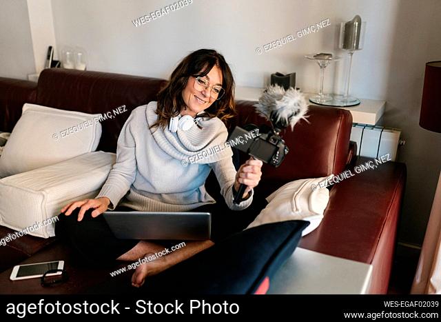 Smiling female vlogger with laptop using camera while vlogging at home