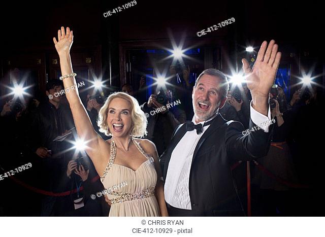 Celebrity couple waving on red carpet with paparazzi in background