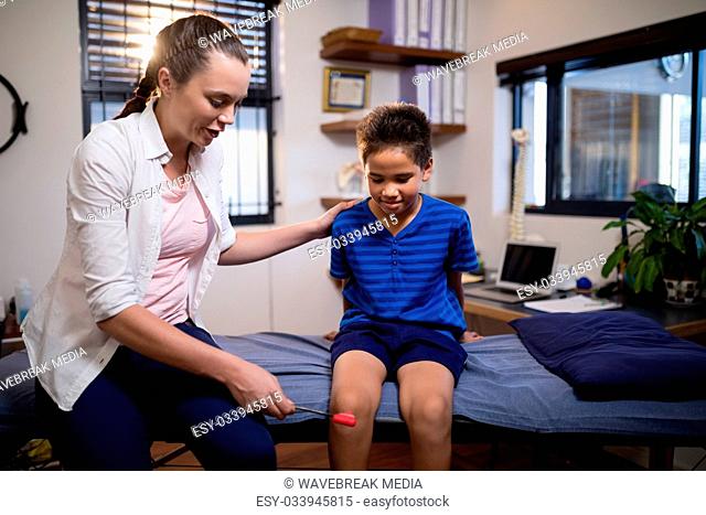 Young female therapist examining boy with reflex hammer on knee while sitting on bed