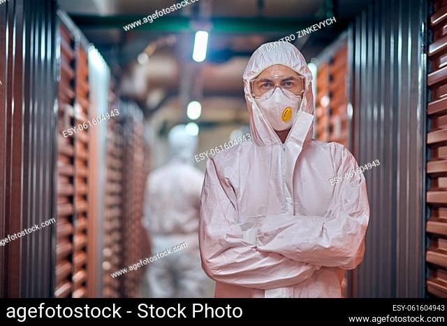 Warehouse employee in a respirator mask and hazmat suit standing with his coworker in the aisle between storage containers
