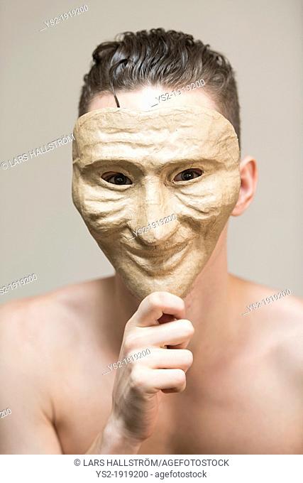 Portrait of a fragile and undressed young adult man hiding behind a happy mask