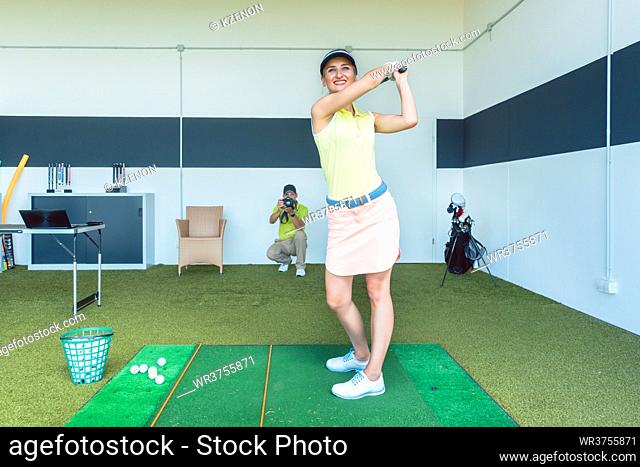 Full length of an attractive fit young woman practicing golf swing while being video recorded by her instructor during professional class indoors