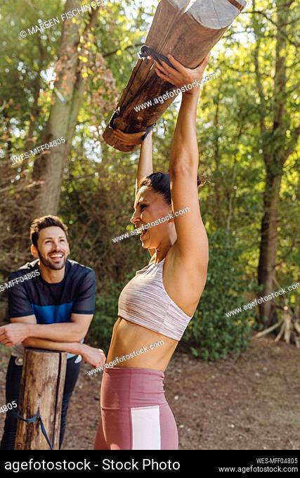 Man watching happy woman lifting up heavy logs on a fitness trail