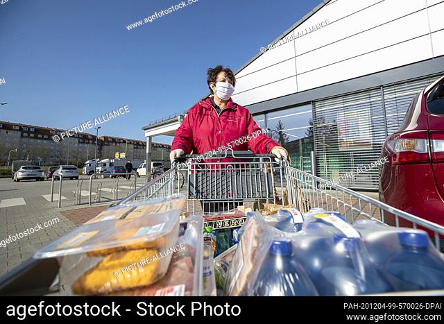 24 March 2020, Saxony, Heidenau: A woman wears a mouth-and-nose protector and disposable gloves and pushes the purchases to the car in a parking lot