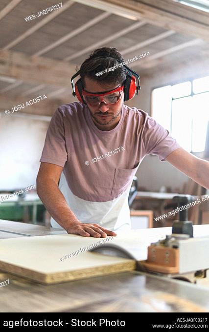 Young male carpenter cutting wood using table saw while working in workshop